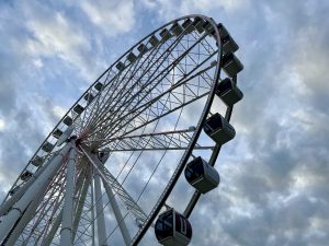 A white Ferris Wheel with multi-colored lights against a blue sky filled with fluffy white clouds.
