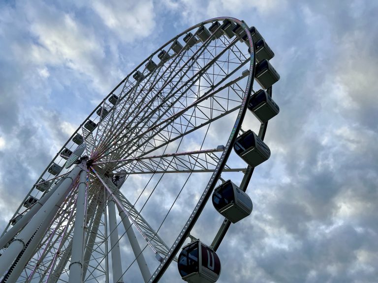 A white Ferris Wheel with multi-colored lights against a blue sky filled with fluffy white clouds.