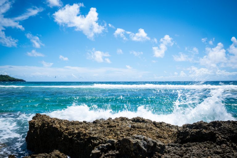 Ocean view, wave crashing up over a rock in the foreground.