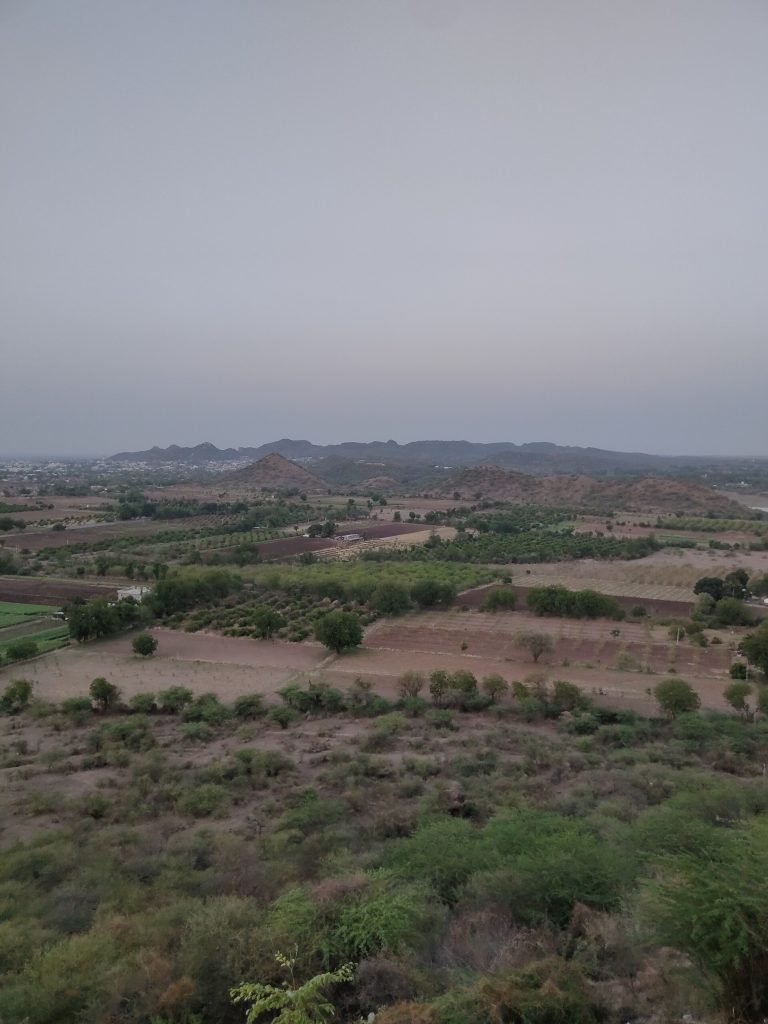 A vista view of fields, some are fields of prepared soil some contain trees and crops, the area looks very dry and dusty. In the background to the left there are lots of buildings and beyond those and to the left is a hilly area.