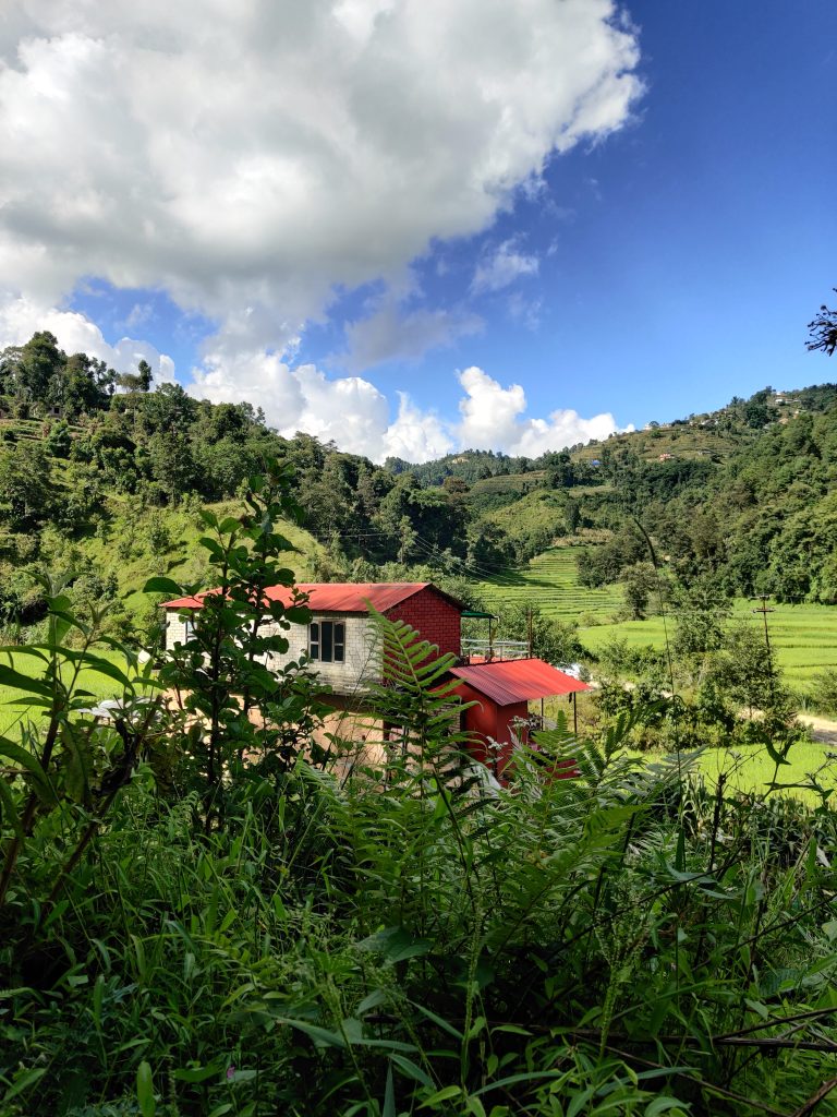 Small, simple brick cabin with balcony and red tin roof surrounded by grass, trees and ferns. Cabin has a matching annex to the right. Houses can be seen in the distance on the hillside.