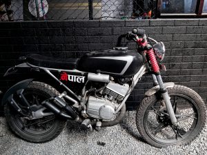 Motorbike side view, leaning against a black brick wall with a black chain link fence above the wall and a building behind the fence.