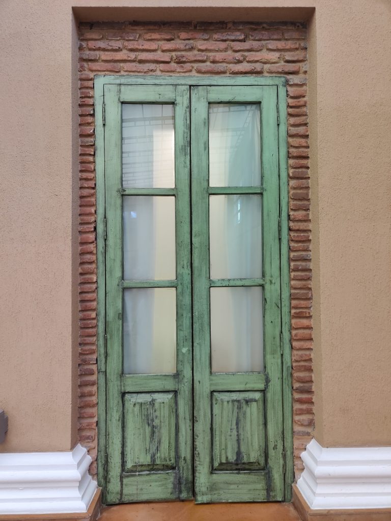 A green double door with glass panels set in a brick wall, framed by white pillars