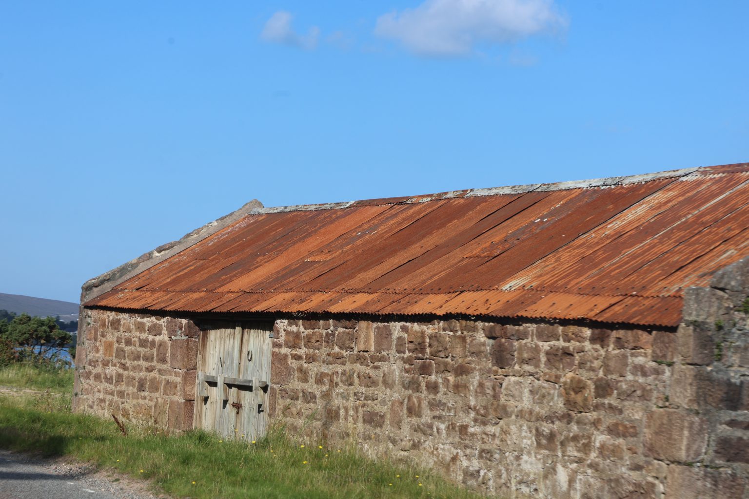 An old stone barn with a rusted tin roof and weathered wooden doors under a clear blue sky.