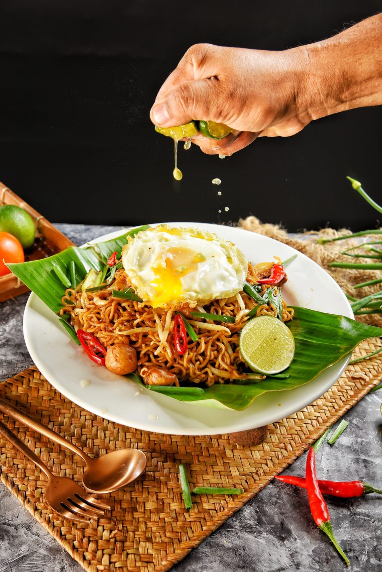 Maggi Goreng, a plate with a banana leaf on it with noodles (maggi) mixed with peppers.  A fried egg on top. A hand is above the plate squeezing a lime with lime juice squirting down.