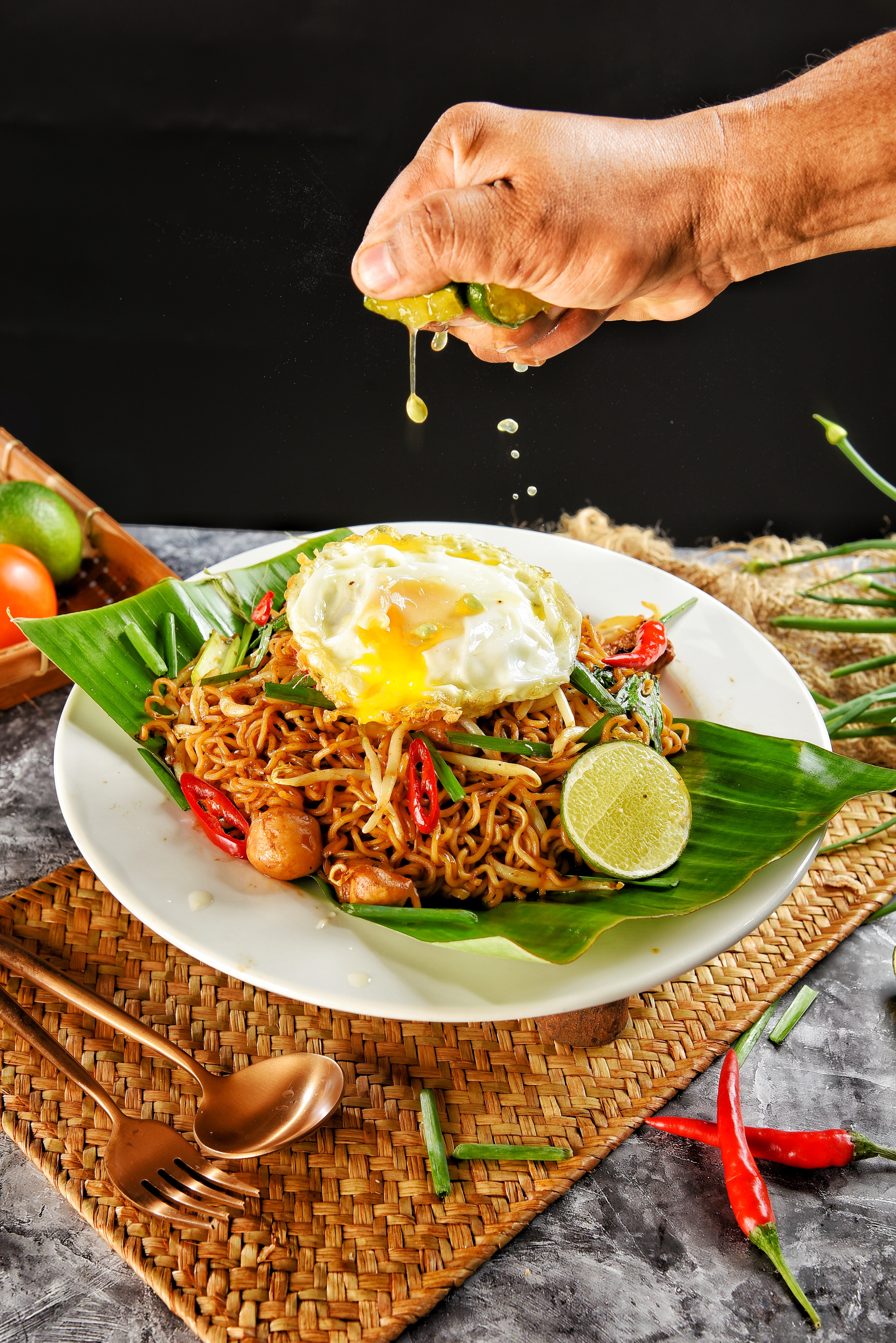 Maggi Goreng, a plate with a banana leaf on it with noodles (maggi) mixed with peppers. A fried egg on top. A hand is above the plate squeezing a lime with lime juice squirting down.