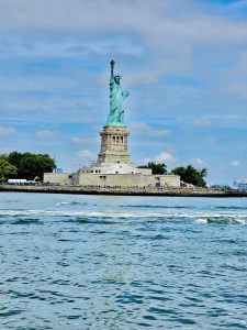 A long view of Statue of Liberty, New York Harbor, United States, taken during a Hudson River cruise. Lots of people beneath the statue and several skyscraper buildings to the right. Scaffolding at the rear of the statue.