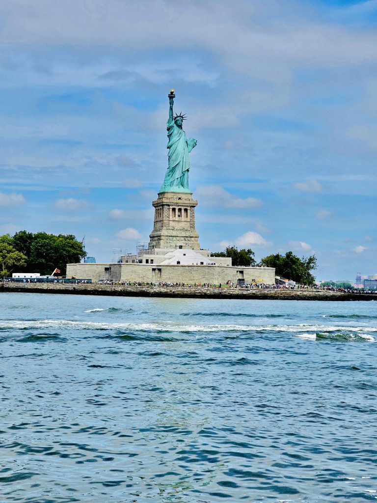 A long view of Statue of Liberty, New York Harbor, United States, taken during a Hudson River cruise. Lots of people beneath the statue and several skyscraper buildings to the right. Scaffolding at the rear of the statue.