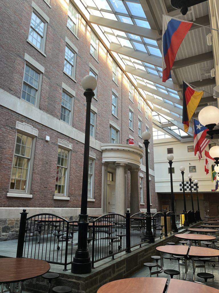 Common area at the Rochester School of the Arts.  Interior that has one wall that looks like a brick exterior wall.  Room has a glass ceiling.  National flags from around the world near the ceiling.