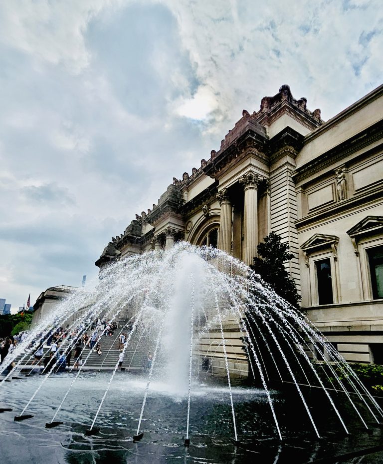 A water fountain in front of the Metropolitan Museum, New York, United States. Lots of people sitting on the musemen steps, skyscrapers can be seen in the distance.