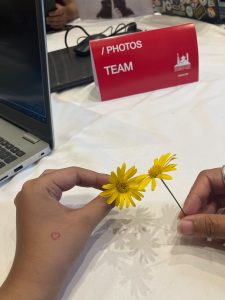 Two hands holding yellow flowers. A sign says "photos team" on the table.