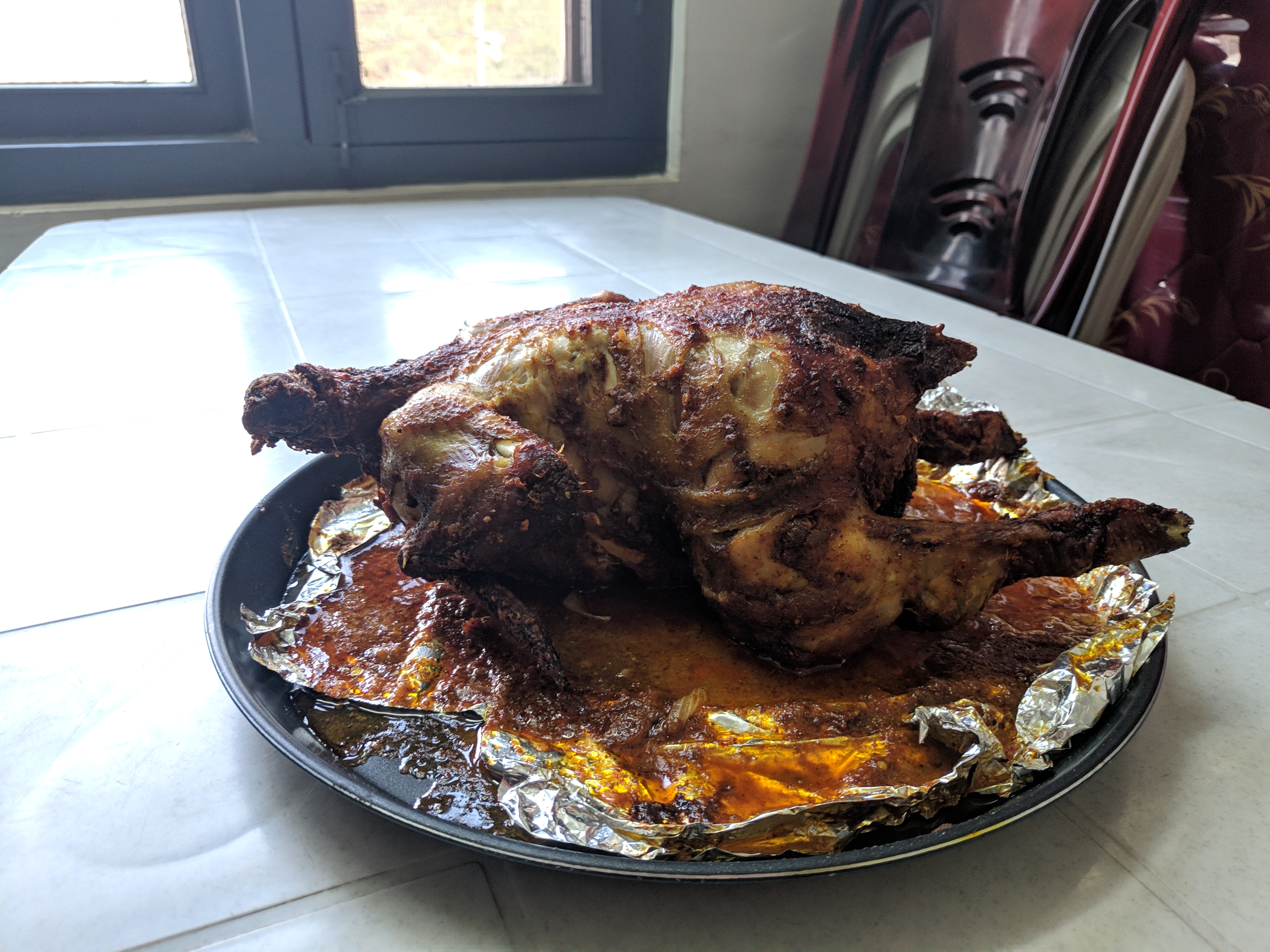 Food, a cooked chicken ready to eat, sitting on aluminium foil and a circular tray, placed on a tiled table beside a window.