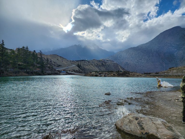 View of mountains, trees, and cloudy sky from the lake. Sunlight is blocked by clouds.