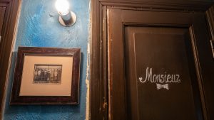 Men’s room toilet, wooden door painted brown with hand painted sign ‘Monsieur’ in white paint with a bowtie painted below it. Textured wall painted blue. A wall mounted light to the left of the door with lit bulb without a lampshade. Below the light it an old photograph in a brown wooden frame, showing a row of small seems boys facing the wall with their backs to the camera as though using a urinal.
