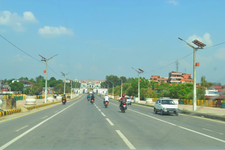 Singha Durbar Palace, Kathmandu, street scene with motorbikes riding away into the distance towards the palace, cars travelling away from the palace on the right hand side, the road is flanked by buildings, solar powered street lights and trees.