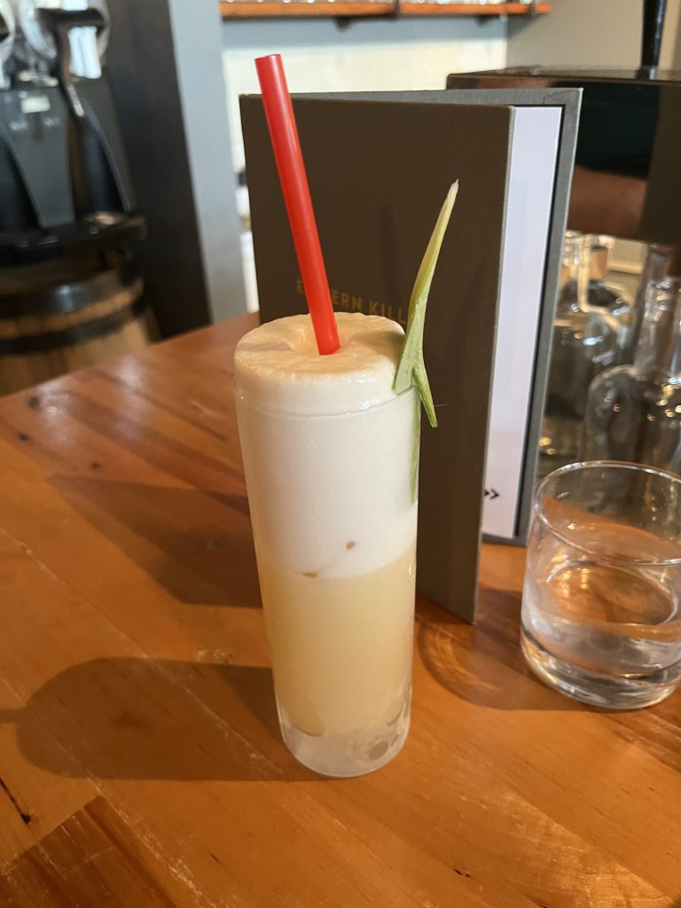 Tall slender cocktail with the foam at the top rising above the edge of the glass, garnished with a pineapple leaf.