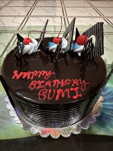A glazed chocolate birthday cake, decorated with cherries, chocolate shards and confectioner’s cream with greeting piped red lettering.
