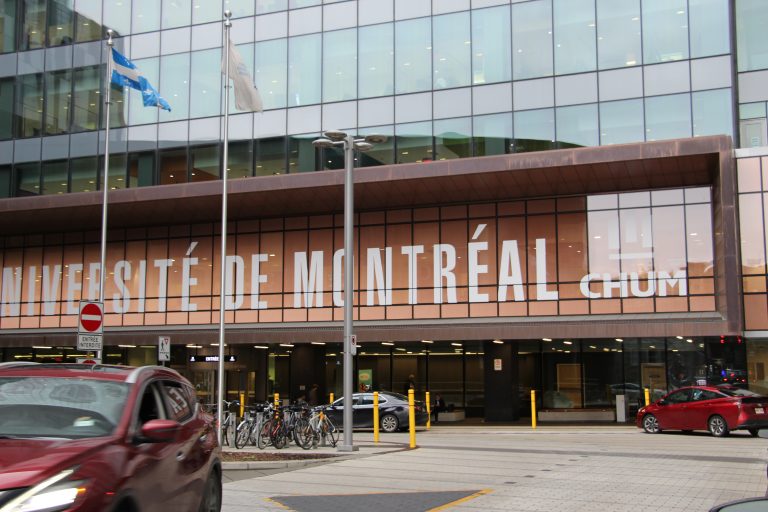 The front entrance of CHUM a university in Montreal, Canada. The building is mainly a glass construction. In the foreground there are bicycles, flag poles and cars, bollards, signposts and paving.