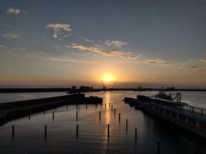 View larger photo: A sunset over a harbor. The sun casts a golden hue on the water. There's a dock in the foreground with a boat alongside it. Beyond the dock are barriers and industrial structures. The sky has scattered clouds, and the horizon meets the sea.