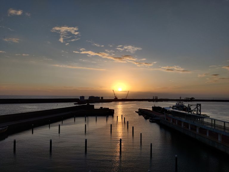 A sunset over a harbor. The sun casts a golden hue on the water. There’s a dock in the foreground with a boat alongside it. Beyond the dock are barriers and industrial structures. The sky has scattered clouds, and the horizon meets the sea.