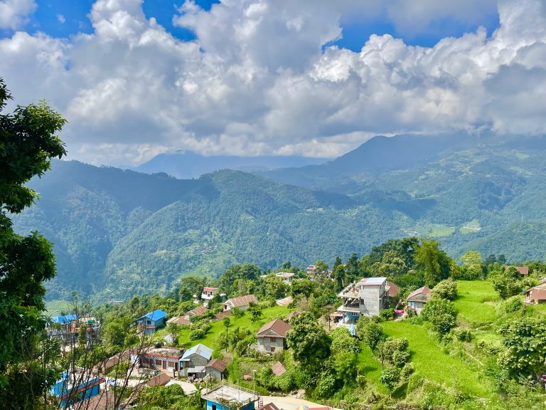 A long view of a Pokhara valley village from Pumdikot hills, Kaski district, Nepal. The picture shows hill top houses amongst the trees, with a blue sky and white clouds in the backdrop.