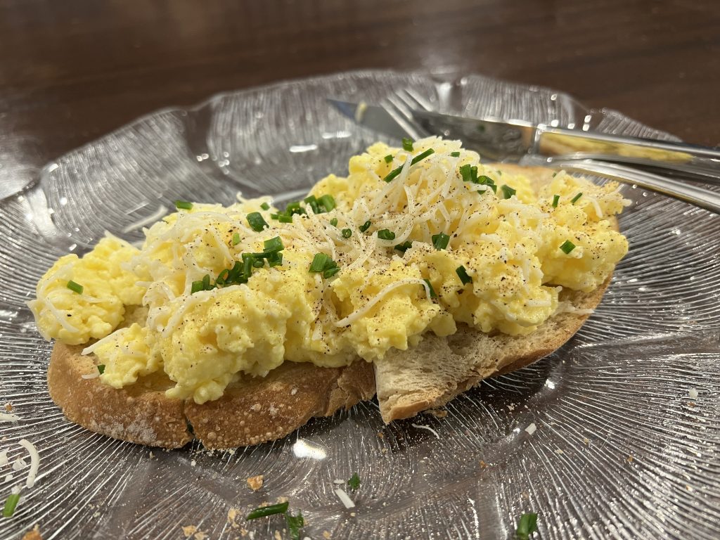 Scrambled eggs with shredded cheese and chopped chives atop toast on a clear glass plate.