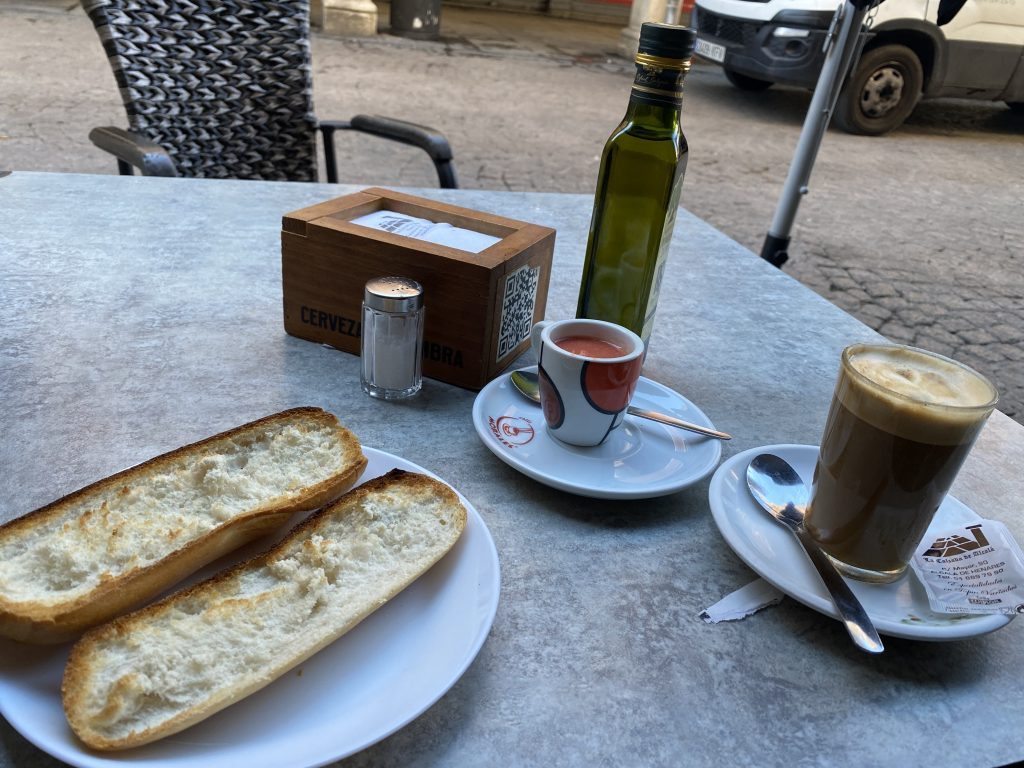 A bar terrace table on the street with a coffee, bread, natural tomato and a bottle of olive oil