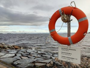 A vibrant orange lifebuoy hangs on a metal pole by a rocky shoreline, with turbulent waters stretching out to a distant horizon dotted with islands. Below the lifebuoy, a sign reads "Nallikarin Uimaranta - Nallikari Beach" and provides emergency numbers. Overhead, the sky is cloudy.