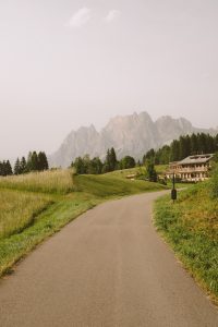 Open sidewalk opening up to a house surrounded by forest trees and mountains looming in the background. 