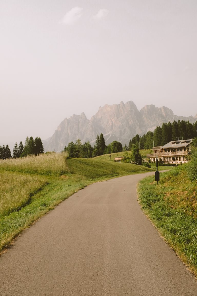 Open sidewalk opening up to a house surrounded by forest trees and mountains looming in the background.