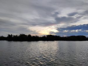 A body of water with ripples on its surface. On one side, there's a line of dense vegetation. Above, the sky is filled with various shades of clouds, with hints of sunlight peeking through, suggesting either a sunrise or sunset.