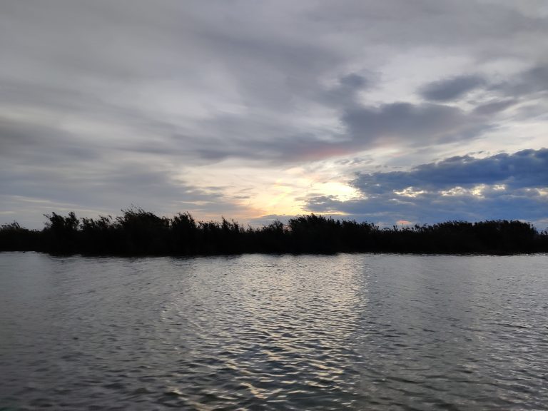 A body of water with ripples on its surface. On one side, there’s a line of dense vegetation. Above, the sky is filled with various shades of clouds, with hints of sunlight peeking through, suggesting either a sunrise or sunset.