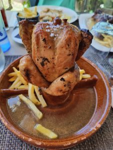 A close-up photo of a roasted chicken seasoned with salt on a brown plate. There are fries on the plate and other dishes in the background. The table has a woven texture. There's a glass and a bottle in the background.