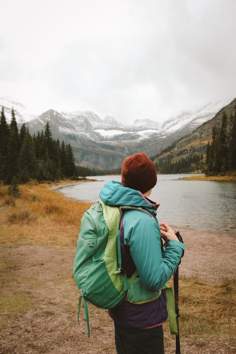 Person in hiking gear and a backpack looking out onto a snowy, fall mountain view with a lake in the foreground.