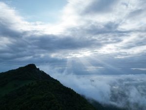 Sunbeams shining down through clouds onto mist and fog in a valley beyond a mountain. 