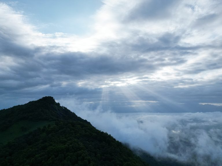 Sunbeams shining down through clouds onto mist and fog in a valley beyond a mountain.