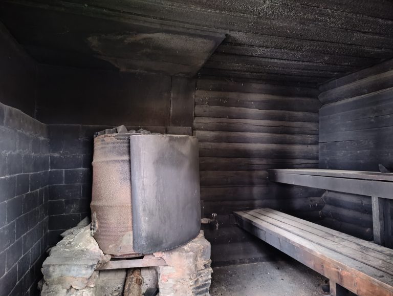 A dimly lit, rustic interior features a large metal stove, rested on a stone foundation, against a blackened wall. The room’s walls are made of dark, aged wooden planks. To the right, wooden benches form a seating area, all hinting at a traditional sauna or smokehouse ambiance.