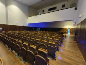 An empty auditorium with rows of brown seats, blue aisle lights, and a wooden floor. The walls are white with a section of brown paneling. There's an exit door on the right side.