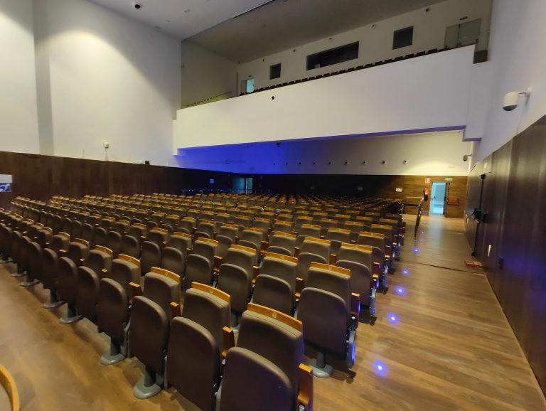 An empty auditorium with rows of brown seats, blue aisle lights, and a wooden floor. The walls are white with a section of brown paneling. There’s an exit door on the right side.