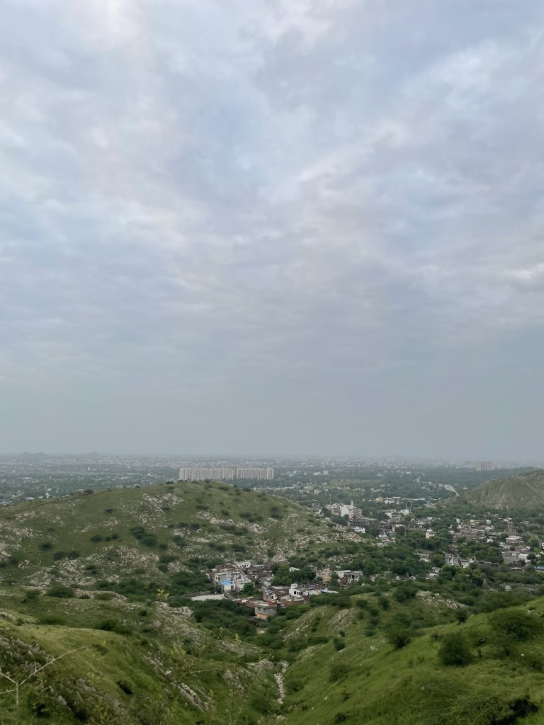 A panoramic view of Jaipur from a mountain peak, featuring a clear blue sky, lush greenery, and the cityscape of Jaipur spread out below. (#WCBhopal)