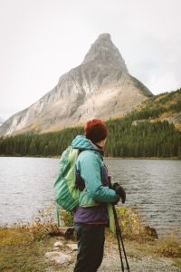 Person wearing winter gear looking back on a view of a lake in front of a triangular shaped mountain surrounded by forest trees. 