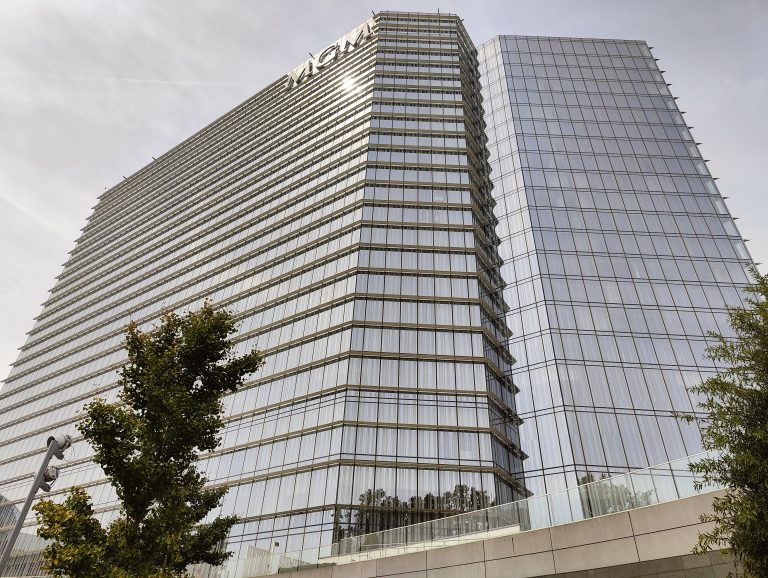 Two tall glass buildings with horizontal window patterns. In the foreground, there’s a tree and a security camera mounted on a pole. The sky is overcast.