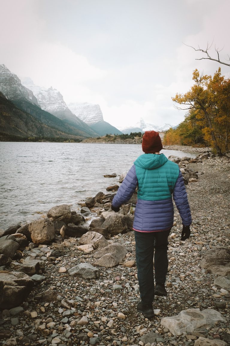 Person in winter hiking gear walking along a rocky shoreline with mountains in view.