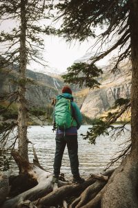 Person in winter hiking gear standing between two small trees looking out over a lake with tall mountains and a waterfall in the background.