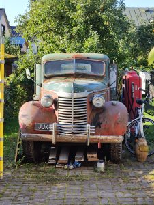 An old rusted truck parked on a paved surface, with items stored beneath it. A red bicycle leans against the truck. Trees with green leaves and apples are behind. Nearby is a yellow pole and a house.