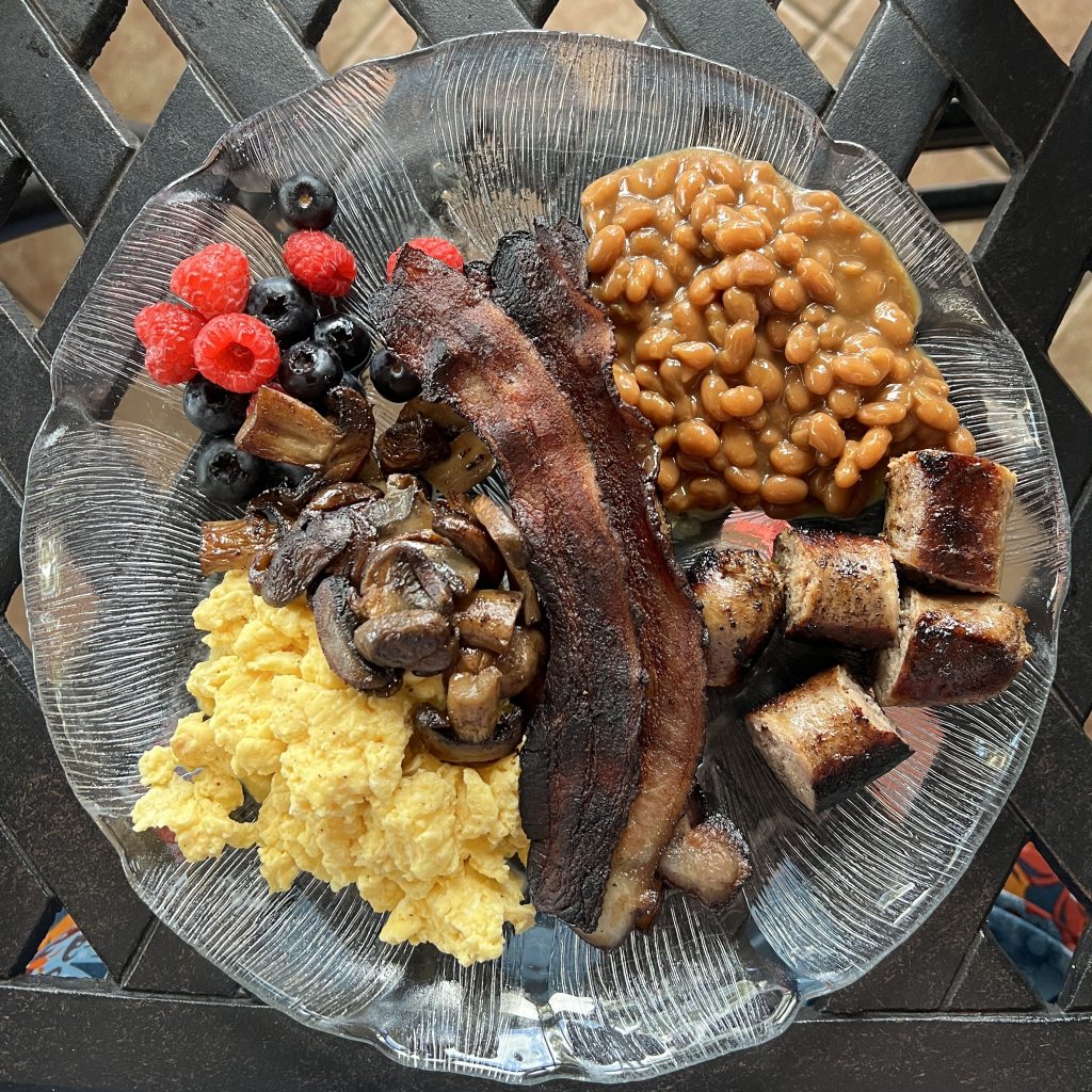 A breakfast plate with mixed berries, scrambled eggs, sausage, bacon, beans, and mushrooms.