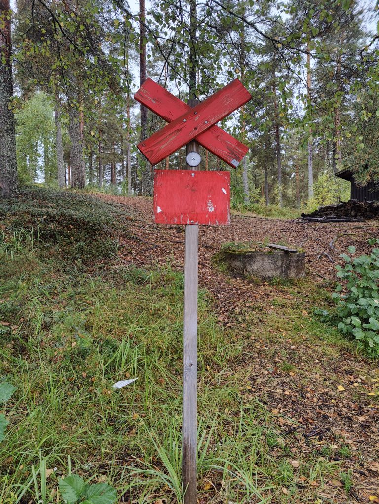 A worn red wooden signpost, with two intersecting planks, stands prominently in a forest clearing. Behind it, tall pine trees rise, their trunks surrounded by green and brown undergrowth. A small wooden structure is partially visible in the background. A white feather lies on the ground nearby.