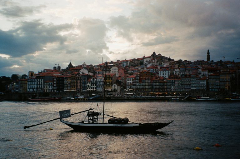 View of the city of Porto from the banks of the Douro River at dusk, with a Rabelos, a traditional local boat, in the foreground on the water.