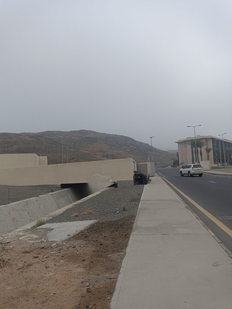 Sidewalk leading away from the camera, street on the right with a small SUV in the road, water runoff channel on the left.