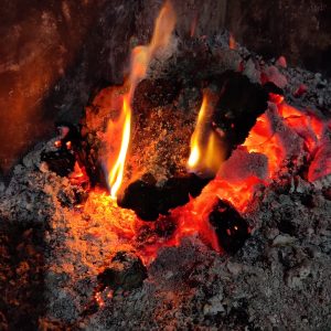 A close-up of burning embers and flames. The embers are glowing and surrounded by ash. There are visible flames, emerging from the embers. The background contains charred wood and ash.
